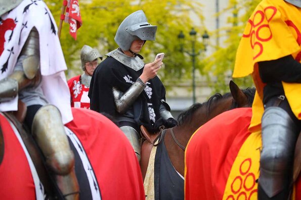 Participants en costumes médiévaux montés sur des chevaux décorés de couleurs vives, lors d'une célébration des fêtes de Jeanne d'Arc. Le cavalier au centre consulte son smartphone.