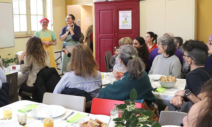 La photo montre un groupe de personnes rassemblées dans une salle lumineuse de la cantine solidaire. Au centre de l'image, deux personnes se tiennent debout, faisant face à l'assemblée. La personne à gauche porte un tablier jaune et un bandana rose, tandis que la personne à droite est vêtue d'un tablier bleu et d'un bandana coloré. Elles semblent s'adresser au groupe, peut-être pour donner des instructions ou partager des informations.  Les participants sont assis autour de tables recouvertes de nappes blanches, sur lesquelles sont disposés des assiettes, des verres et des fleurs. Les personnes présentes semblent attentives et engagées dans la conversation. L'ambiance est conviviale et communautaire, suggérant un espace où les gens se réunissent pour partager un repas et échanger. À l'arrière-plan, une porte rouge est visible, avec une affiche sur le mur à côté.