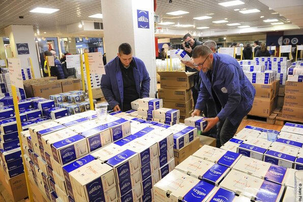 Scène animée à l'intérieur de la boutique Duralex d'Orléans, avec deux employés portant des blouses bleues. Ils manipulent des boîtes empilées de produits Duralex, principalement des verres en carton d'emballage bleu et blanc