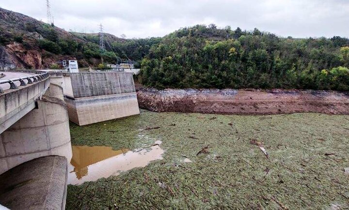 Vue d'un barrage en béton avec une structure en arc à gauche, situé dans un environnement naturel avec une colline boisée en arrière-plan. À l'avant-plan, une grande étendue d'eau recouverte de débris flottants, principalement des végétaux et des branches, après une crue de la Loire. Le ciel est nuageux, et des pylônes électriques sont visibles sur la colline