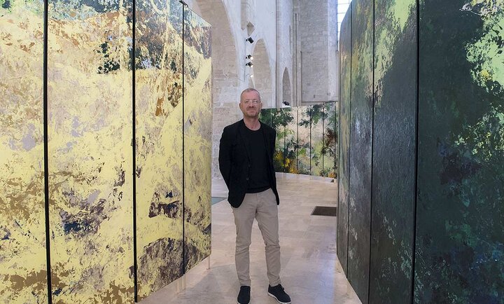 Sur cette photo, Bruno Desplanques, vêtu d’une tenue sobre composée d’un blazer noir, d’un t-shirt assorti et d’un pantalon beige, se tient debout au milieu d’une installation artistique immersive. L’espace, baigné de lumière naturelle, est une vaste salle aux hautes voûtes en pierre, évoquant l’architecture d’un édifice historique.  Autour de lui, de grands panneaux peints, aux couleurs et textures abstraites, créent une atmosphère entre nature et abstraction. Les tons oscillent entre des nuances de vert profond, de jaune doré et des touches plus sombres, conférant à l’ensemble une impression de paysage fragmenté. L’installation semble transformer l’espace en une œuvre vivante, où les visiteurs peuvent circuler et se laisser envelopper par les jeux de couleurs et de matières.  Le lieu, la collégiale Saint-Pierre-le-Puellier d’Orléans, offre un contraste saisissant entre l’art contemporain et la pierre ancienne, soulignant la rencontre entre patrimoine et création artistique moderne.