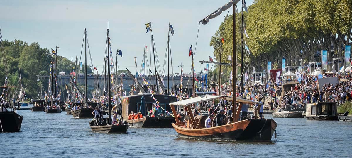 la grande parade pavoisée de clôture du Festival de Loire à Orléans. Sur la Loire, un grand nombre de bateaux traditionnels en bois – futreaux, toues et gabares – voguent en cortège, richement décorés de drapeaux, fanions et bannières colorées. Des mariniers, certains en costume d'époque, saluent la foule ou manœuvrent lentement. La berge droite est noire de monde : spectateurs massés sous les arbres, le long des quais ornés de guirlandes et de stands. En arrière-plan, le pont George V est également envahi par le public venu admirer ce moment fort du festival. L’ambiance est festive, patrimoniale et conviviale, portée par la lumière dorée d’une fin d’après-midi estivale.