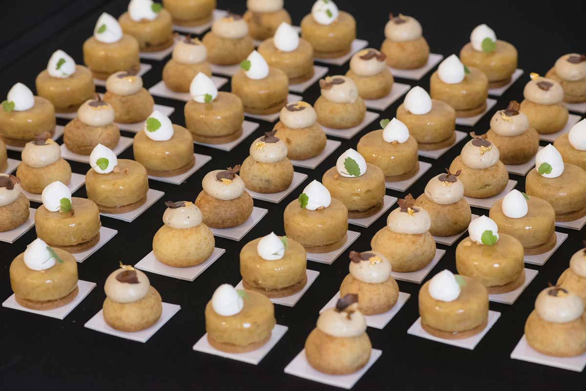 L'image montre une vue en plongée d'une grande quantité de petites pâtisseries individuelles, disposées en rangées serrées sur une surface noire. Chaque pâtisserie est composée de deux parties : une base ronde et plate, surmontée d'une sphère plus petite. Les deux parties sont de couleur beige clair, avec des nuances légèrement dorées, suggérant une cuisson. Chaque pâtisserie est coiffée d'une petite touche de crème blanche, surmontée d'une minuscule feuille verte. Certaines pâtisseries présentent également une petite décoration supplémentaire de couleur marron foncé sur la crème.